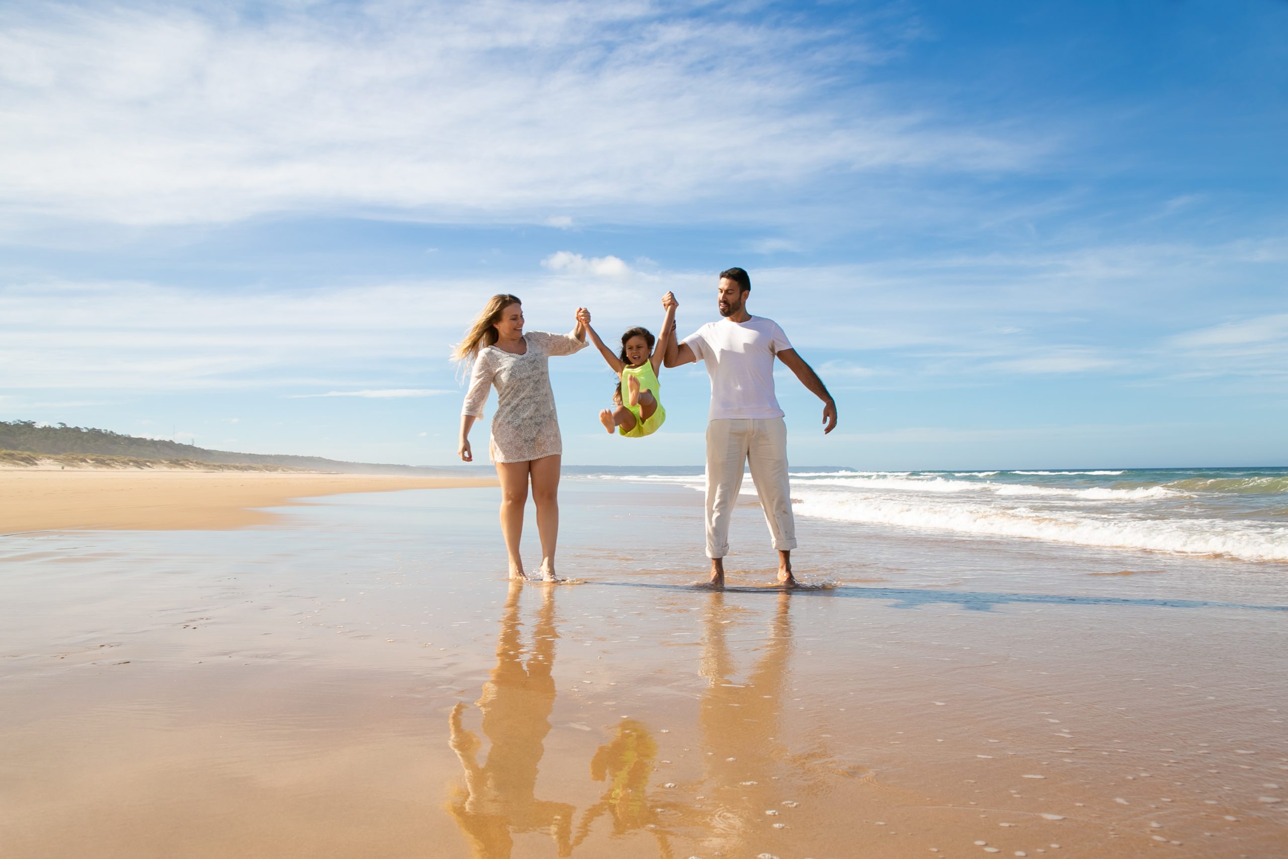 Cheerful parents and little girl enjoying walking and activities on beach, kid holding parents hands, jumping and throwing legs up. Front view. Family outdoor activities concept